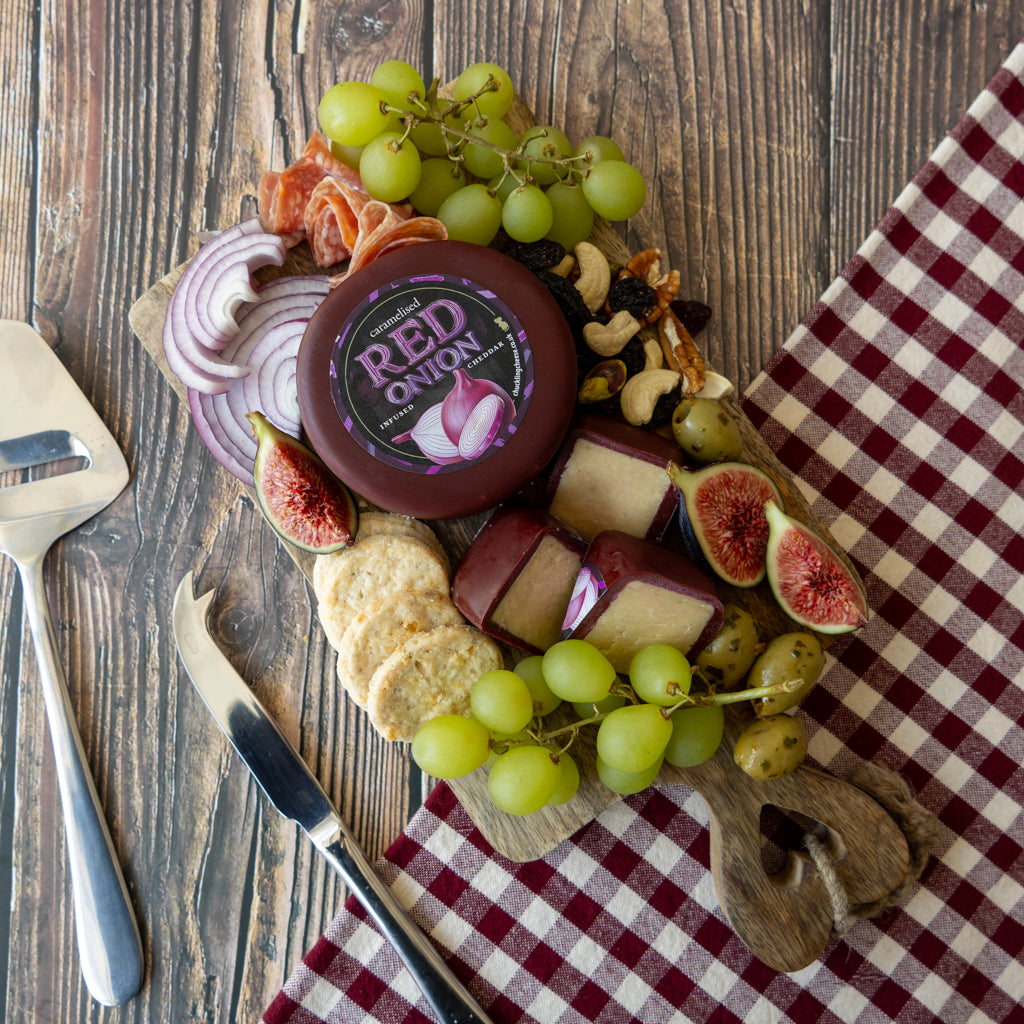 A round wheel of red onion cheese with a dark brown label sits on a wooden charcuterie board surrounded by green grapes, sliced red onion, figs, crackers, cheese wedges, olives, and cured meat. The board is styled on a rustic wooden table with a red-and-white checkered cloth, a cheese knife, and a cheese server.