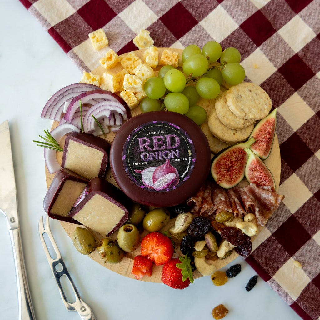 A charcuterie board styled with a round wheel of Red Onion Cheese in the center, surrounded by grapes, sliced red onion, crackers, figs, olives, nuts, dried fruit, and cheese wedges on a wooden board, with a red-and-beige checkered cloth underneath.