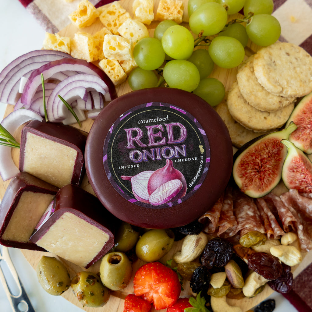 A close-up overhead view of a charcuterie board featuring a round container of caramelised red onion cheddar cheese, sliced and cubed cheddar, green grapes, crackers, figs, olives, strawberries, salami, nuts, and a knife on a white plate.