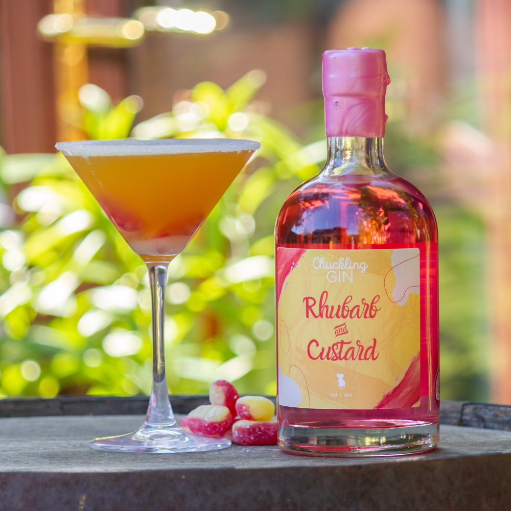 Bottle of Chuckling Gin Rhubarb and Custard with a cocktail and fruit on a wooden surface outdoors.