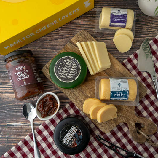 A styled cheese and charcuterie board arrangement on a wooden surface, featuring multiple cheese blocks and slices, a jar of tomato and garlic chutney, a small bowl of chutney, a black container, a wooden cutting board, and a yellow box labelled 'Made for Cheese Lovers'.