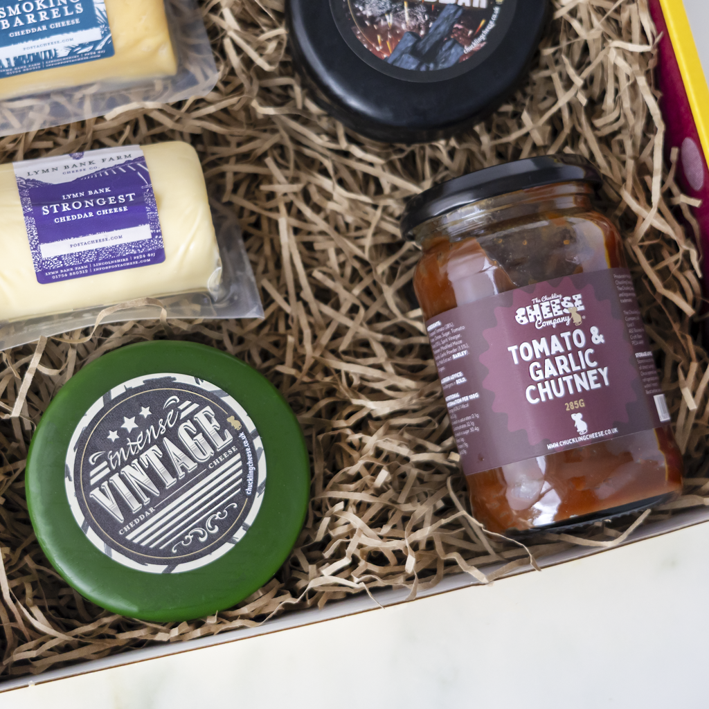 A curated gourmet gift box filled with shredded paper, containing a jar of The Chuckling Cheese Company Tomato & Garlic Chutney, a green round cheese labelled Vintage Creamy Cheese, a packaged cheese block labelled Strongest Cheese, and another round black cheese truckle.