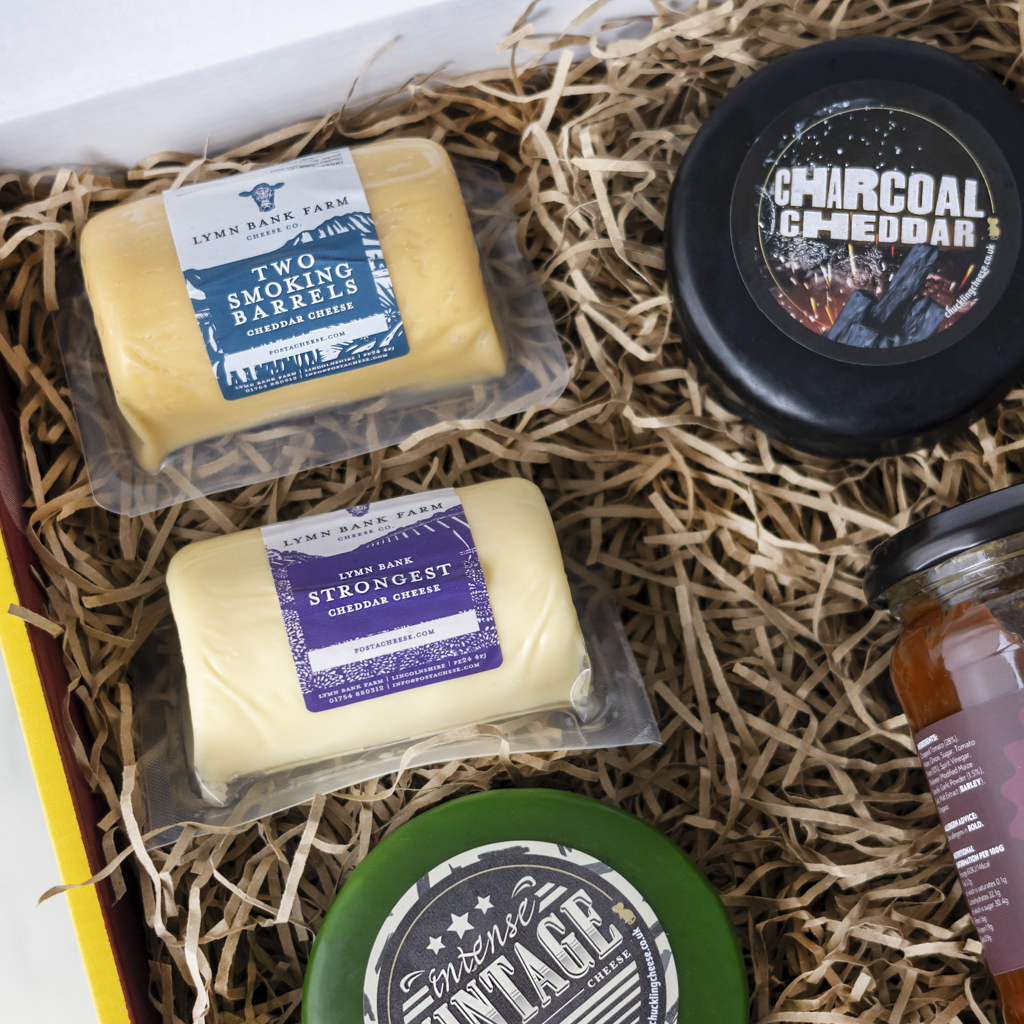 A curated cheese gift box from The Chuckling Cheese Company, featuring two packaged cheddar cheeses, a round charcoal-aged cheddar cheese wheel, a jar of honey, and a green cheese wheel, all arranged in a box filled with brown paper straw.