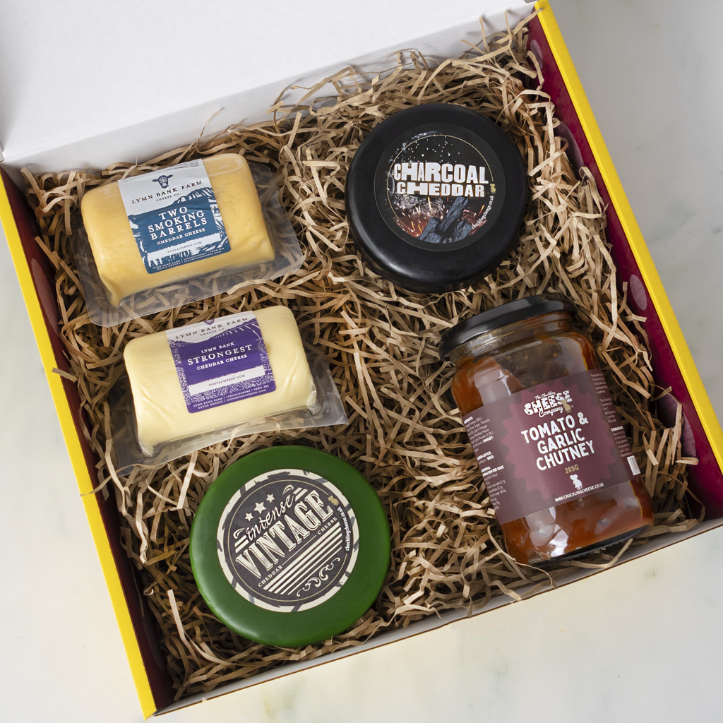 An open gift box filled with shredded paper and gourmet food items, including two packaged cheeses, a black cheese truckle container labelled 'Charcoal Cheddar', a green-lidded jar labelled 'VINTAGE', and a glass jar of 'CHEESE TOMATO & GARLIC CHUTNEY'.