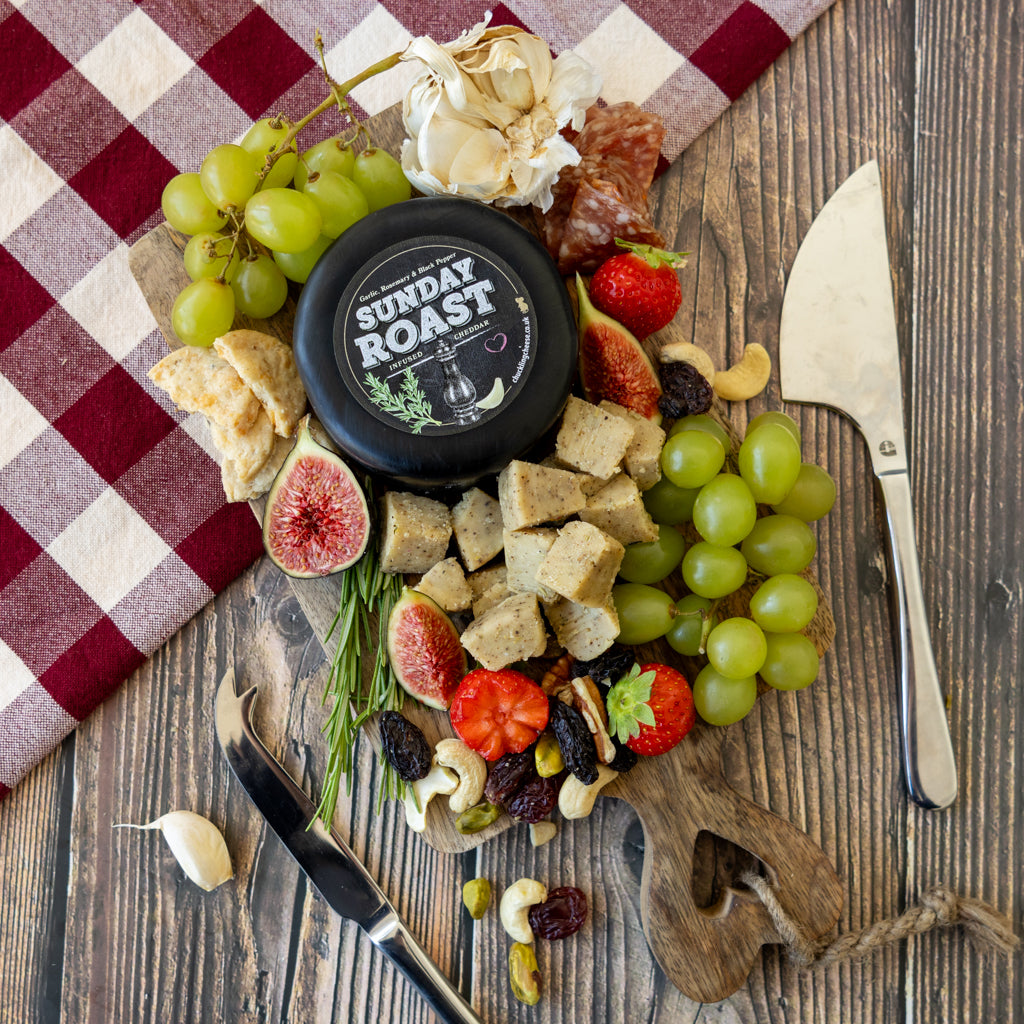 A styled flat-lay of a charcuterie board featuring a round black truckle of Sunday Roast cheese, surrounded by green grapes, figs, strawberries, crackers, nuts, garlic, and a red-and-white checkered cloth on a wooden table.