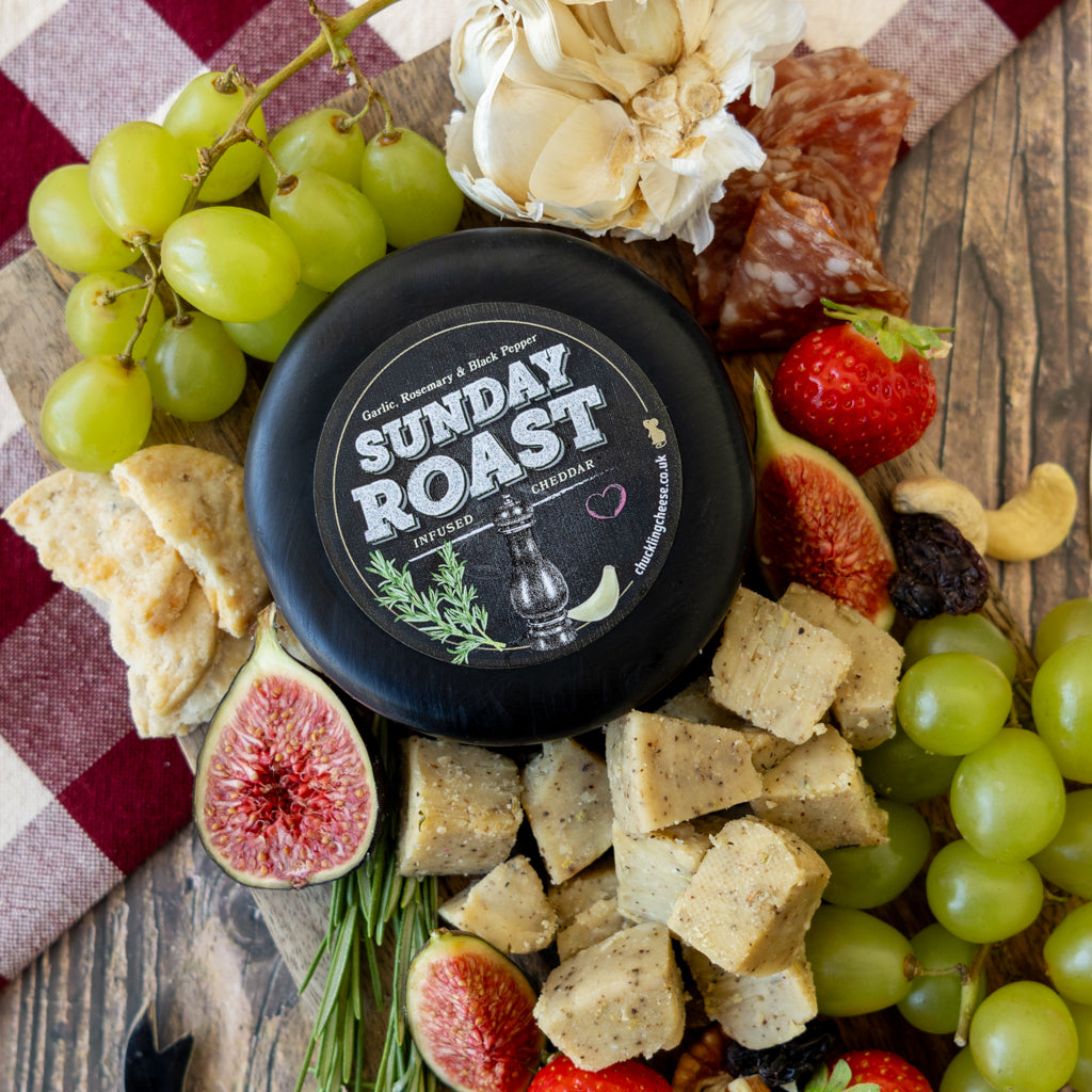 A round black cheese wheel labeled 'SUNDAY ROAST' sits on a rustic wooden board surrounded by green grapes, figs, crackers, garlic, salami, and strawberries.