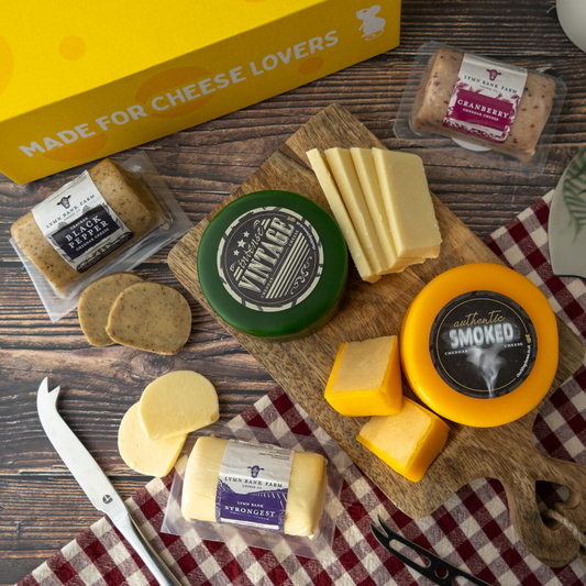 A cheese and charcuterie board arrangement on a dark wood surface, featuring multiple cheeses from The Chuckling Cheese Company, including a green wheel labelled 'Vintage', a yellow smoked wheel, sliced pale cheese, crackers, and a yellow gift box that reads 'MADE FOR CHEESE LOVERS'. A red-and-white checkered cloth and a cheese knife are also visible.