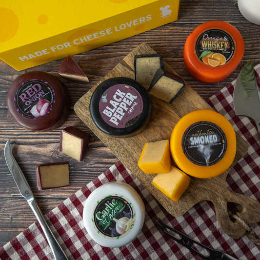 A cheese assortment display with multiple round and cut cheese wheels in different flavors, arranged on a wooden cutting board and table with a yellow 'Made for Cheese Lovers' box, a knife, tongs, and a red-and-white checkered cloth.