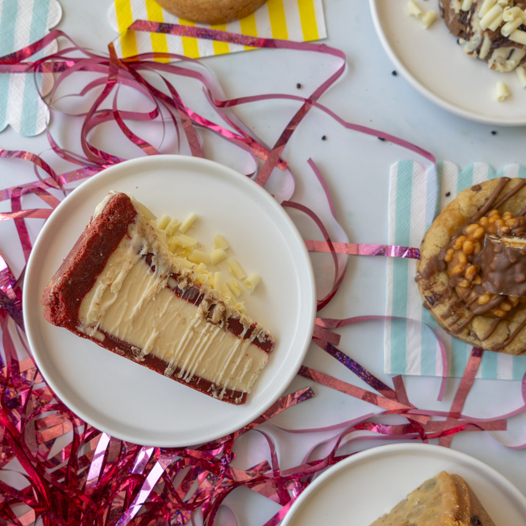 A slice of layered chocolate cake on a white plate, surrounded by pink tinsel ribbon and other dessert slices on plates, arranged on a light tabletop.