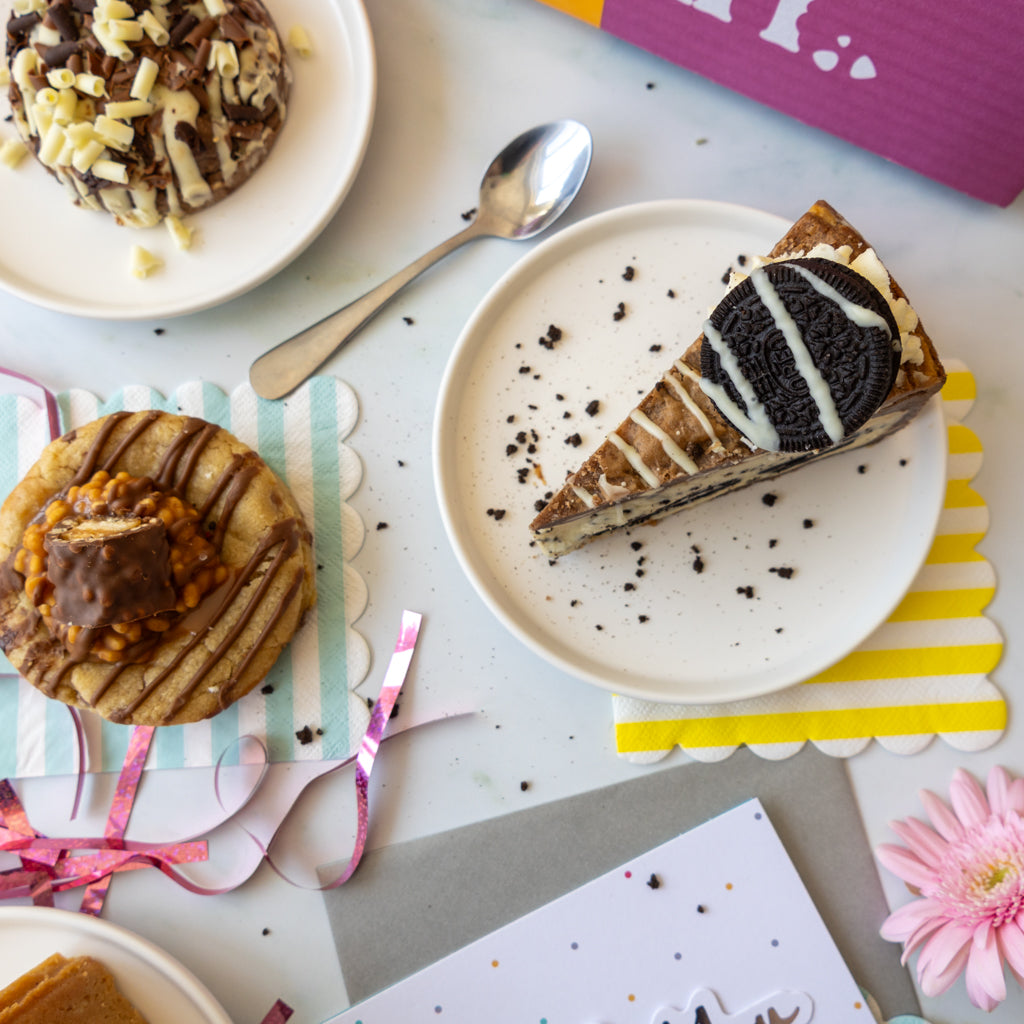 A styled flat-lay of baked goods on a white surface, featuring a slice of chocolate cake with white drizzle and an Oreo cookie on a white plate, alongside two chocolate-covered pastries on white plates, a silver spoon, and colorful paper accents.