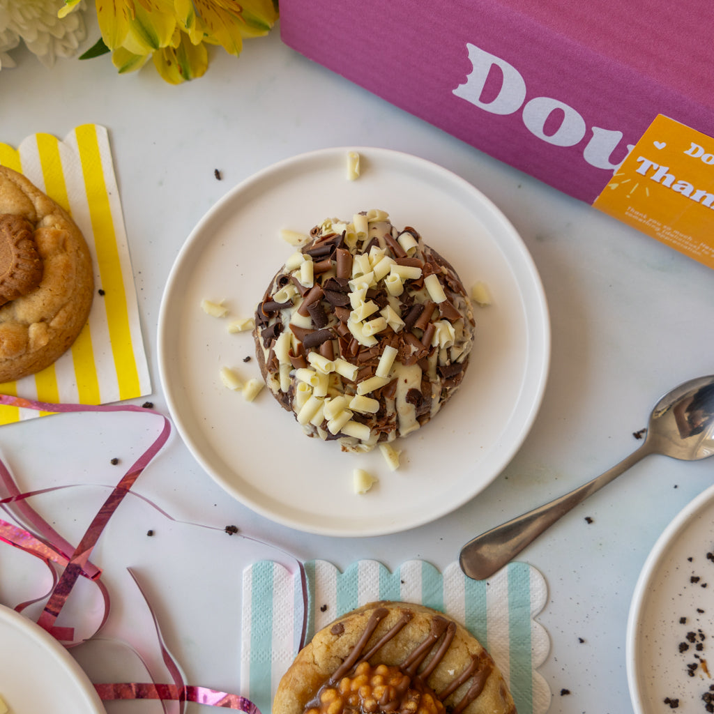 A dessert topped with chocolate and white chocolate shavings sits on a white plate in a styled tabletop scene with pastries, ribbon, a spoon, flowers, and a pink branded box in the background.
