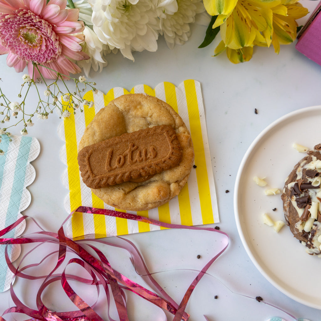 A round cookie topped with a brown biscuit embossed with the word 'Lotus', placed on a yellow-and-white striped napkin on a white surface with flowers, ribbon, and a plate nearby.