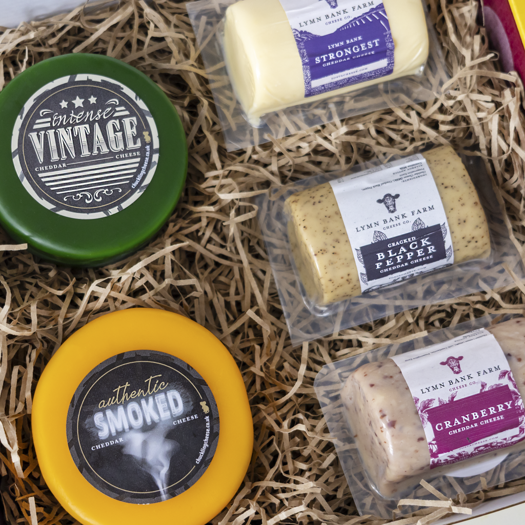 A curated cheese gift box filled with shredded paper, featuring four packaged cheeses from Lymn Bank Farm: an intense vintage semi-hard cheese in a green round container, an authentic smoked cheddar wheel in a yellow container, a black pepper cheese stick, and a cranberry cheese stick.