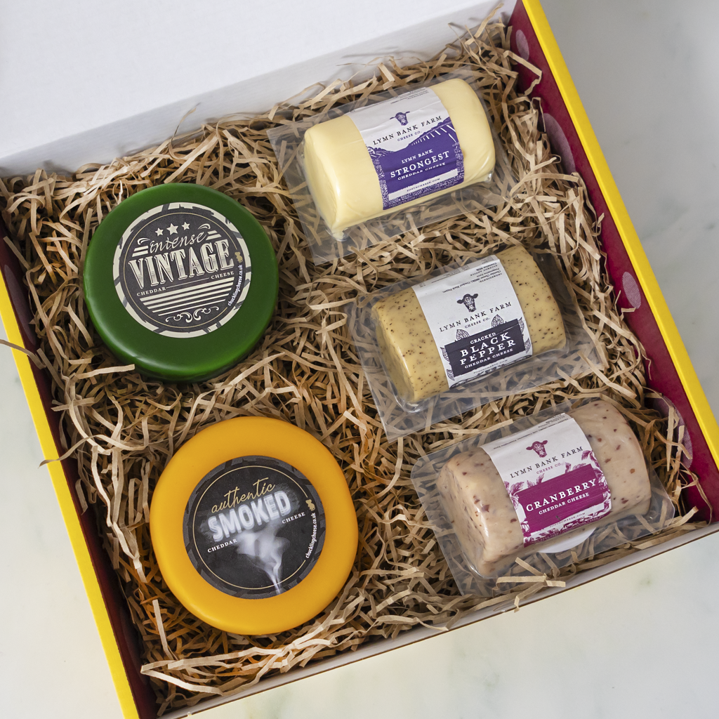 A curated cheese gift box from The Chuckling Cheese Co. filled with shredded paper, containing five different cheeses: a green 'Vintage' round cheese, a yellow 'Smoked' round cheese, a pale yellow 'Strongest' cheese, a beige 'Black Pepper' cheese, and a pinkish-beige 'Cranberry' cheese, all in clear packaging.