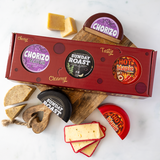 A boxed cheese truckle gift set arranged on a white marble surface with a wooden board. The red box shows three circular flavour labels: Chorizo, Sunday Roast, and Hot Maple.