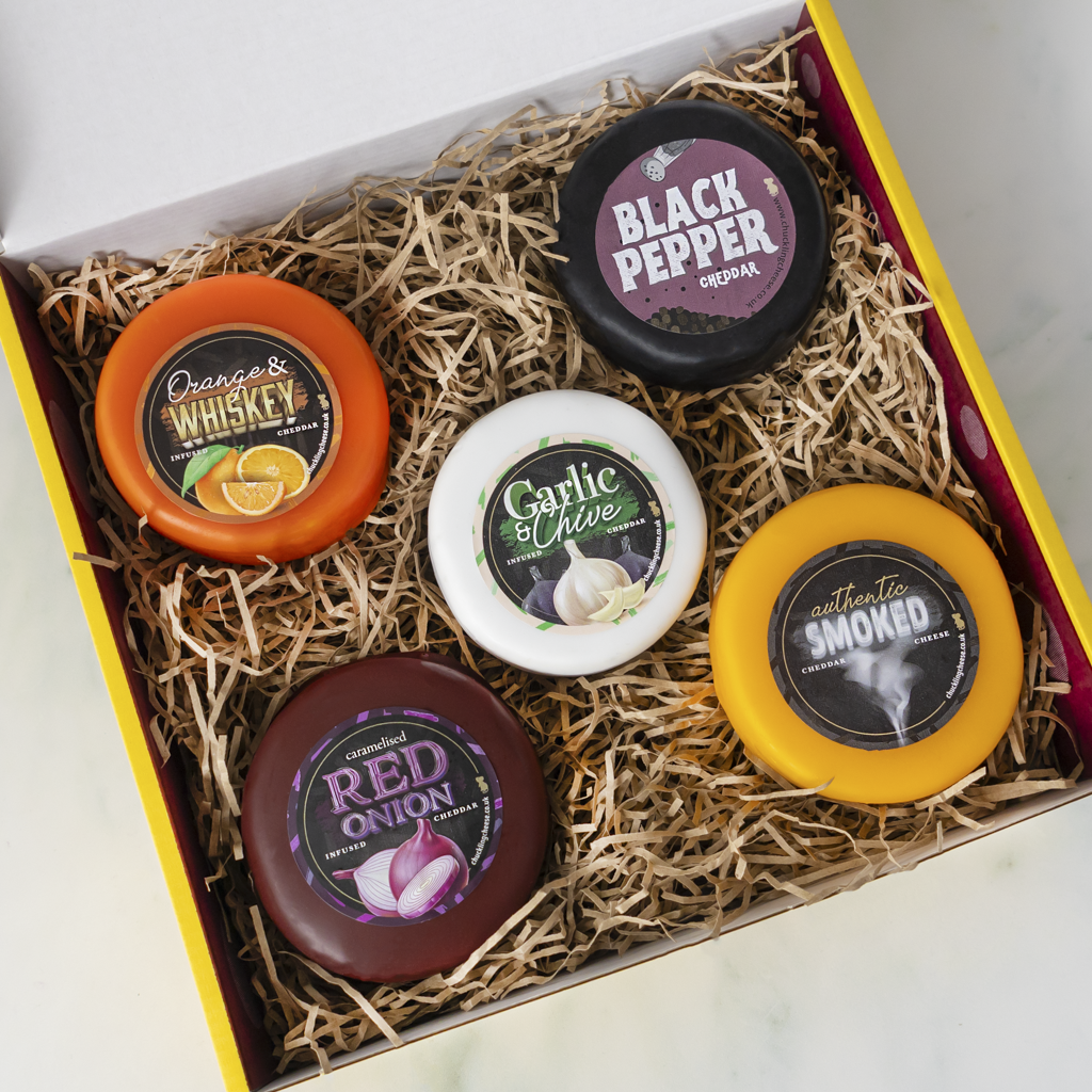 A gift box of five gourmet cheese spreads nestled in shredded paper. The spreads include Orange & Whiskey, Black Pepper, Garlic Olive, Red Onion, and Smoked, each in a round container with a colorful label, set against a light background.
