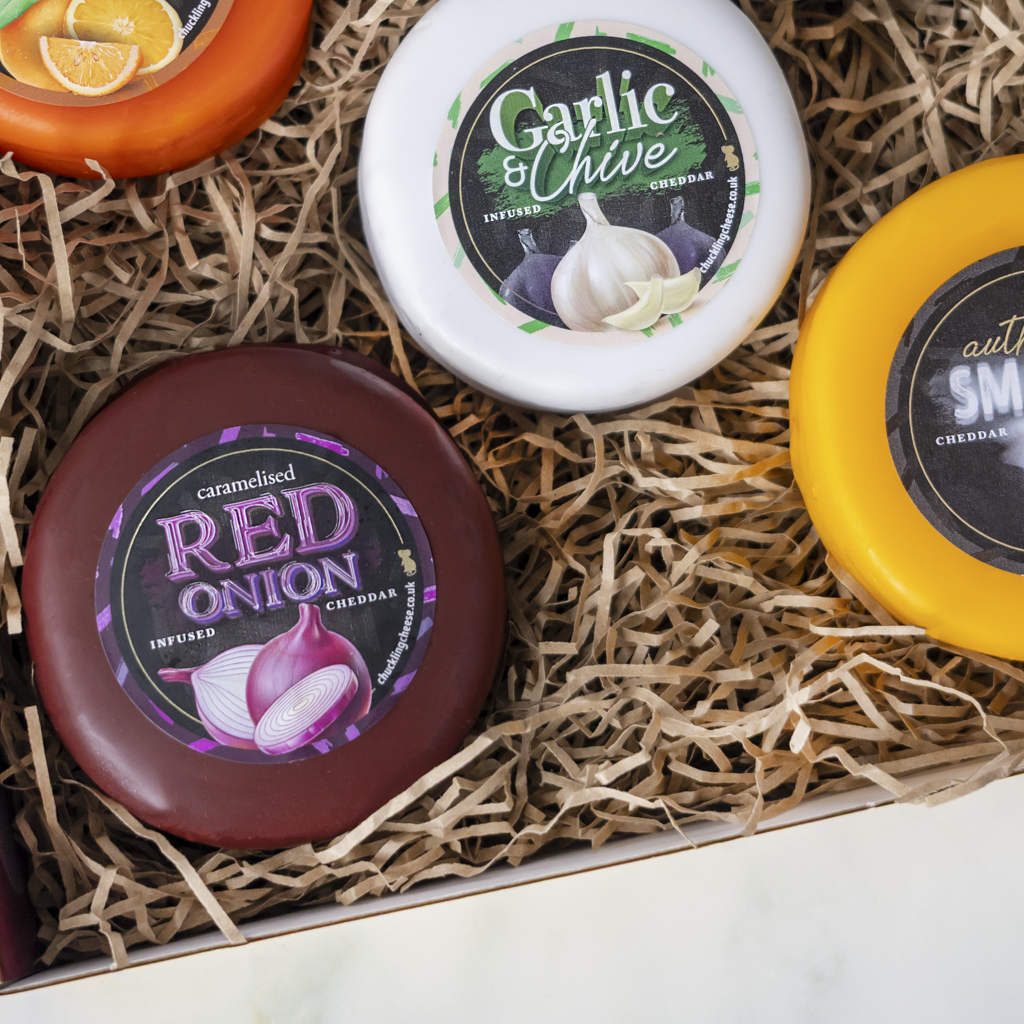 A gift box of infused cheddar cheese wheels nestled in brown paper shred. The visible cheeses include Garlic & Chive, Red Onion, and Smoked, with colorful labels in white, dark red, and yellow.