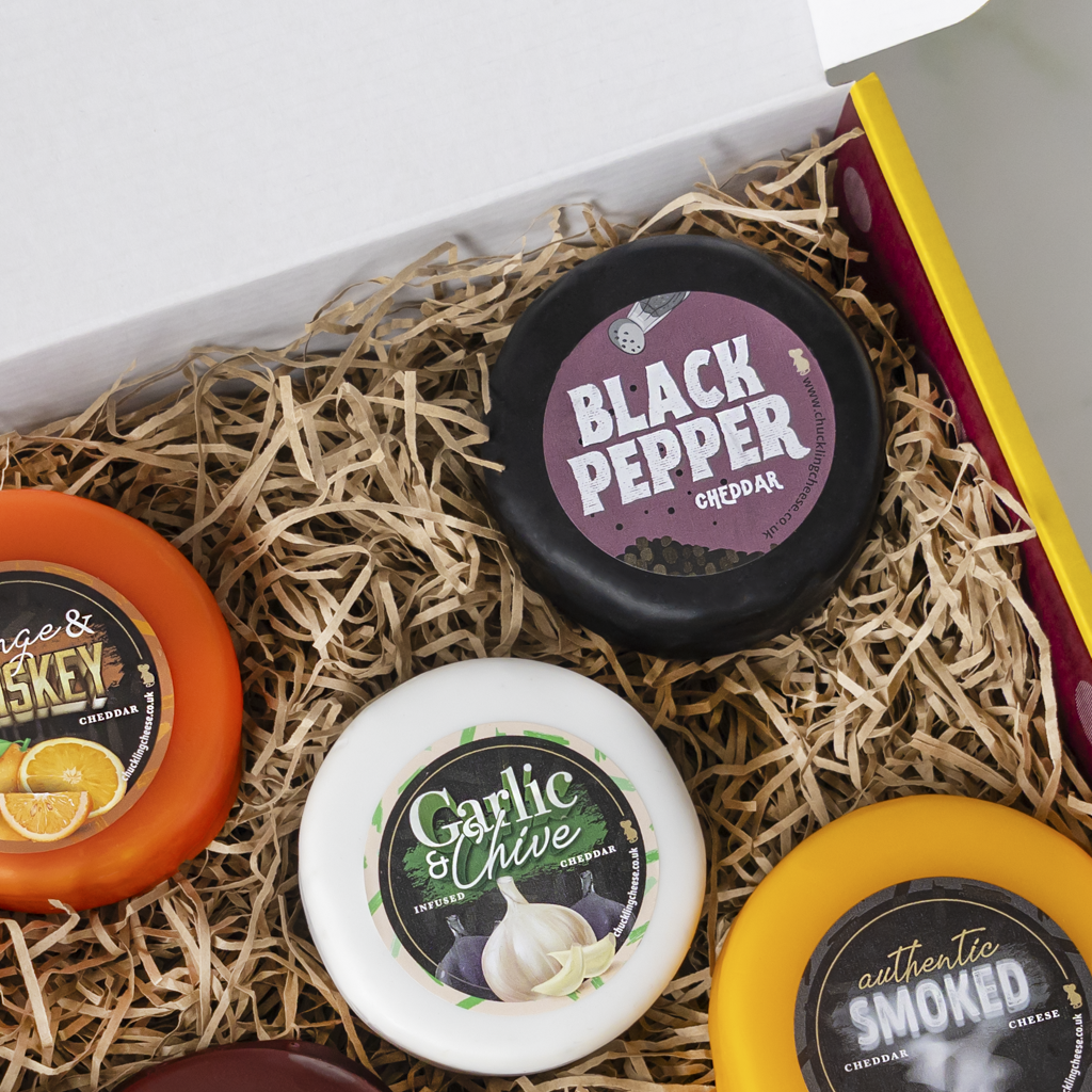 A curated cheese gift box filled with shredded paper, featuring several small round cheddar cheese wheels with colorful labels including Black Pepper, Garlic & Chive, authentic Smoked, and orange and yellow varieties.