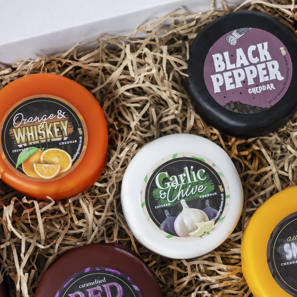 Six small round cheese wheels arranged in shredded straw packaging, showing labels for Orange & Whiskey, Garlic & Chive, Black Pepper Cheddar, Red, and Sharp Cheddar.