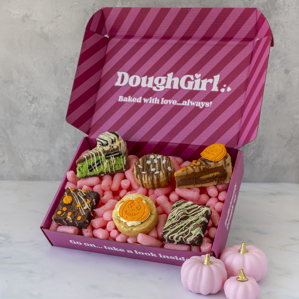An open pink DoughGirl gift box filled with assorted baked goods, and small pink pumpkins, photographed on a light marble surface against a grey wall.