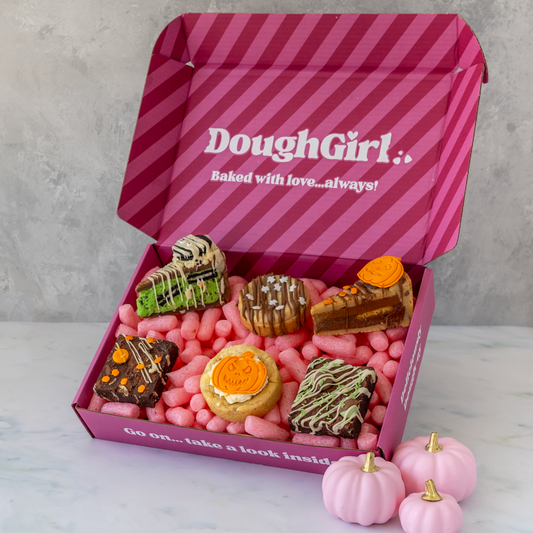 An open pink DoughGirl gift box filled with assorted baked goods, and small pink pumpkins, photographed on a light marble surface against a grey wall.