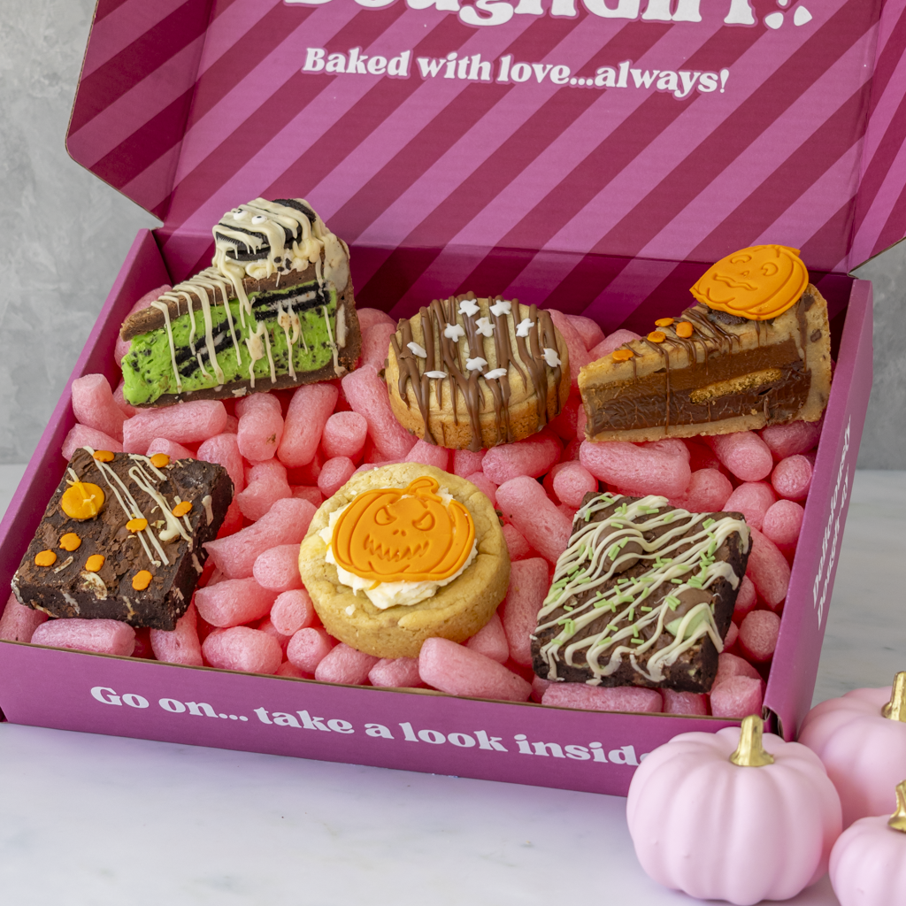 A pink gift box filled with assorted Halloween-themed baked goods, including cookies, brownies, and a slice of cookie pie, with pink candy corn filler inside. The box lid reads 'Baked with love'.