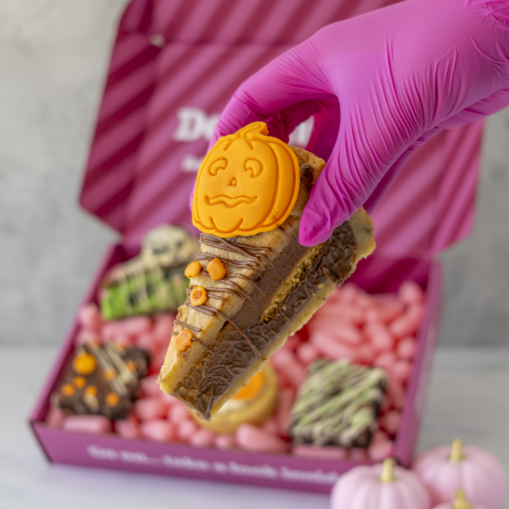 A hand wearing a bright pink glove holds a decorated slice of cookie pie topped with an orange pumpkin fondant. In the background, an open pink gift box filled with pink candies and assorted treats sits on a light surface.