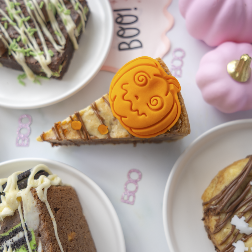 A Halloween-themed food flat lay featuring a slice of cookie pie decorated with a bright orange pumpkin-shaped topper, surrounded by other dessert slices on white plates with BOO-themed props in the background.