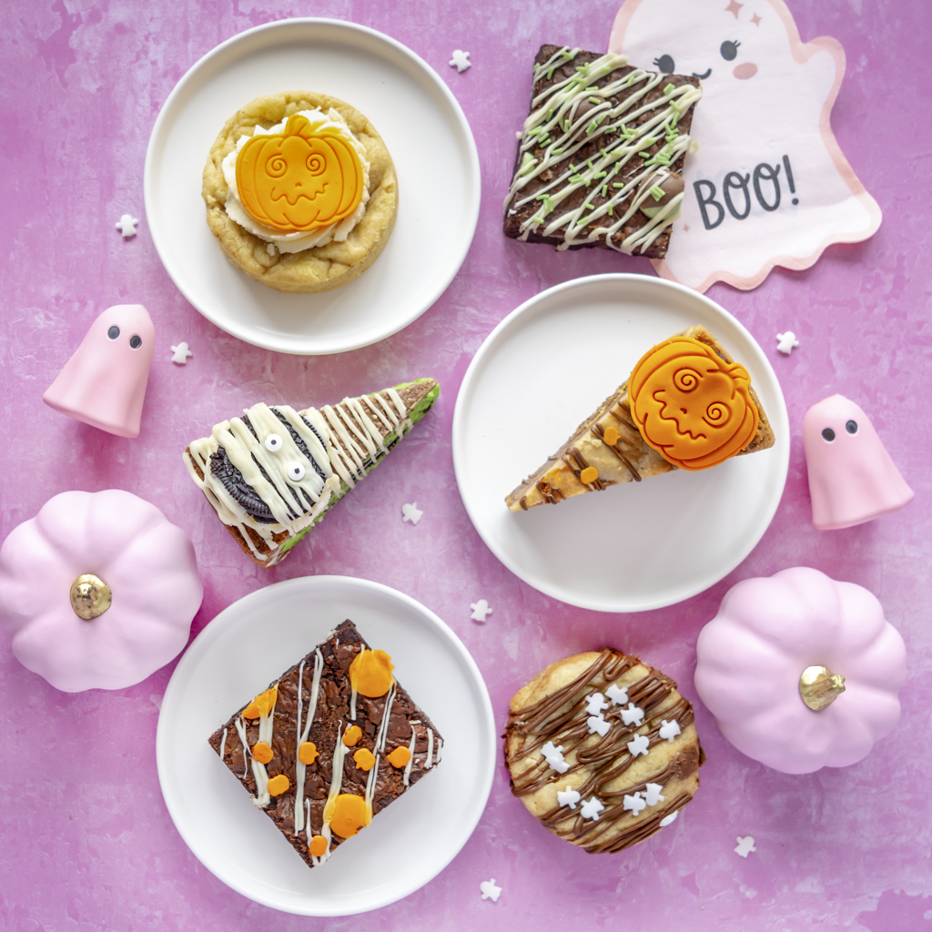 Halloween-themed dessert spread on a pink background with plates of decorated cookies and brownies, plus pink ghost and pumpkin props. Visible treats include a pumpkin cookie, a mummy cake, a ghost cookie, and chocolate drizzle desserts.