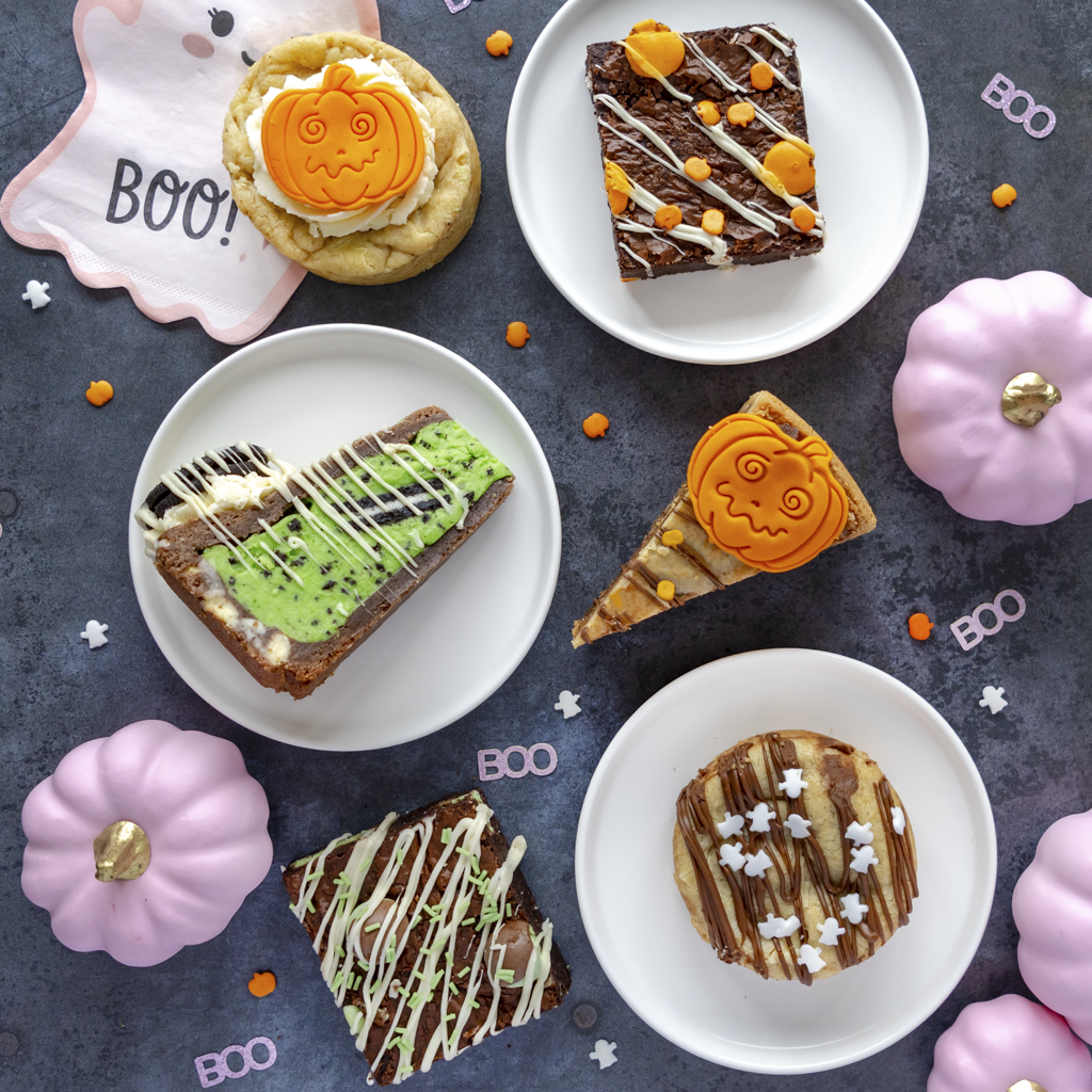 Halloween-themed dessert spread on white plates, featuring pumpkin cookies, chocolate cookie pie slices, and a green pumpkin cookie pie slice, surrounded by pink pumpkins, candy corn, and BOO decorations on a dark textured background.