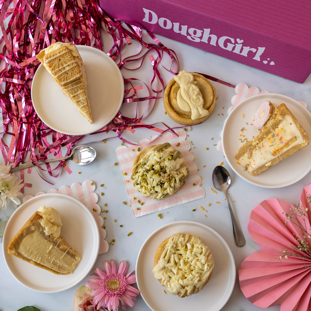 A styled flat-lay of several cookies and pie slices on white plates, arranged with pink ribbon, paper fans, flowers, and a purple box labeled DoughGirl.