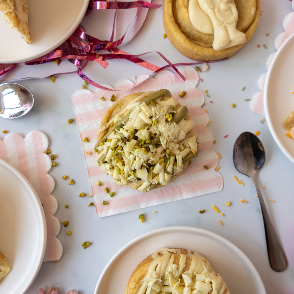 A top-down styled food scene showing a pastry topped with white chocolate and chopped pistachio on a pink-and-white striped napkin, surrounded by plates, spoons, ribbon, and other pastries on a light surface.
