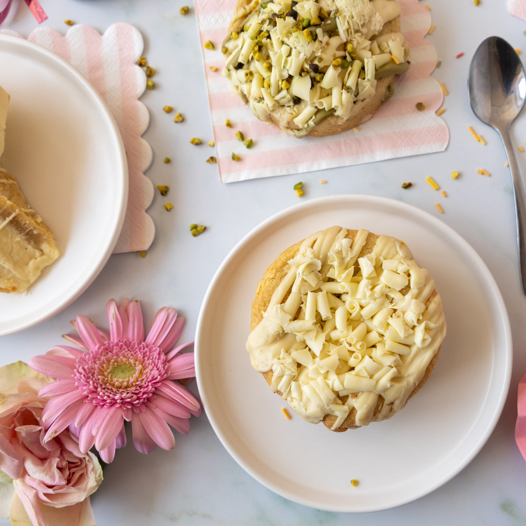 Two small pastries topped with white icing and chopped pistachio, arranged on white plates on a light marble surface with pink flowers, a spoon, and a pink-and-white napkin.