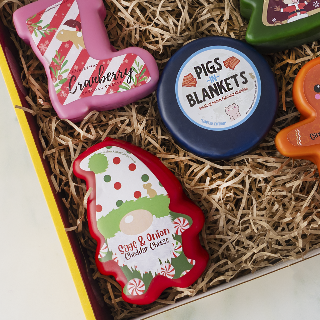 A gift box filled with holiday-themed cheese spreads and snacks, including a pink Christmas stocking spread labelled 'Cranberry Cheddar', a round blue cheese spread labelled 'Pigs in Blankets', a red gnomeshaped spread labelled 'Sage & Onion Cheddar Cheese', and other colourful holiday items in shredded paper packaging.