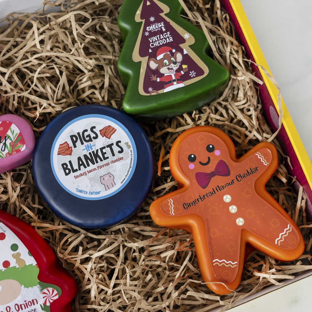 A gift box filled with cheese spreads in decorative shapes, including a gingerbread man, Christmas tree, and round blue tin labeled 'Pigs in Blankets', set in shredded paper packaging.