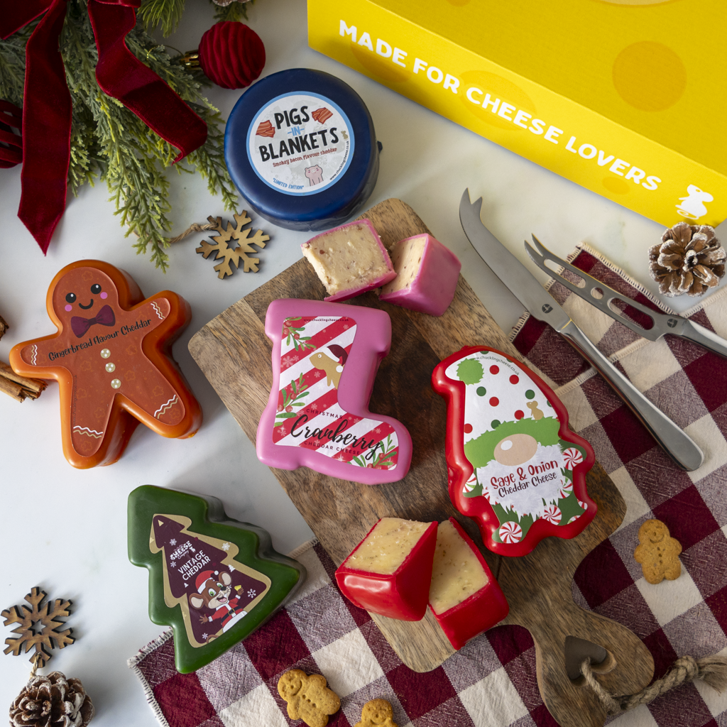 A styled holiday cheese board scene with multiple shaped cheese wheels and blocks, including gingerbread, boot, Christmas tree, and Santa shapes, arranged on a wooden board with carving tools and a yellow 'Made for Cheese Lovers' box on a white surface with festive props.