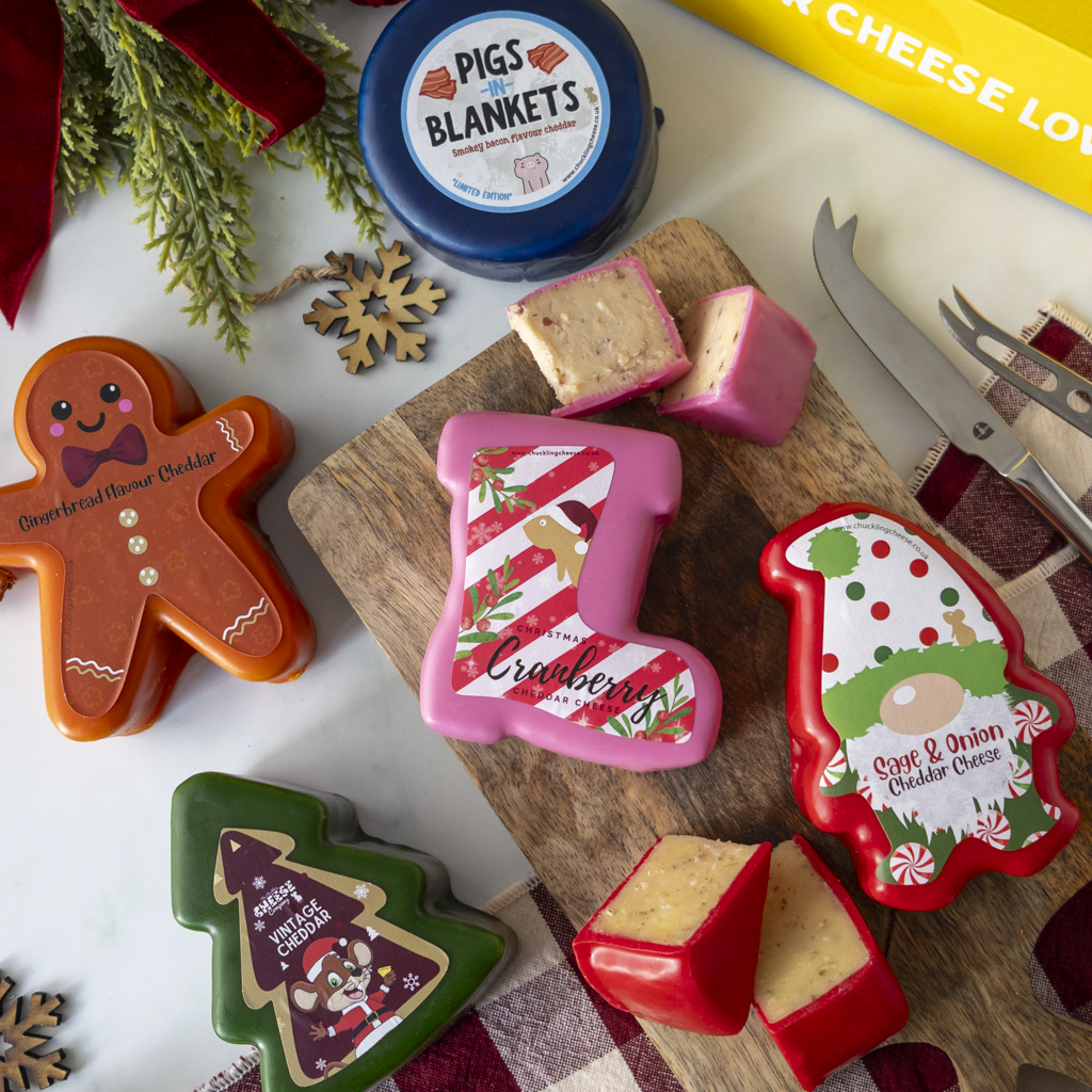 A styled flat-lay of holiday-themed cheese wheels and cheese blocks in decorative tins. The products include gingerbread, Christmas tree, Santa, and Christmas stocking shapes, alongside a blue round tin labeled 'Pigs in Blankets' and a yellow box edge.