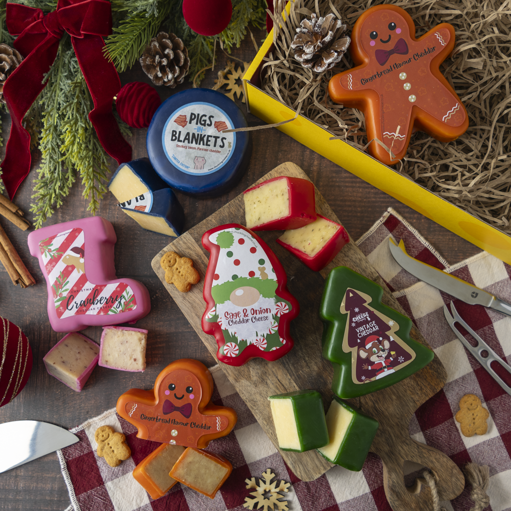 A styled holiday food and gift arrangement on a dark wooden table featuring personalised gingerbread man and Christmas tree cheese wheels with matching cheese wedges, a blue wheel labelled 'Pigs Blankets,' a yellow tray, pinecones, evergreen branches, a red ribbon, and a checkered cloth.