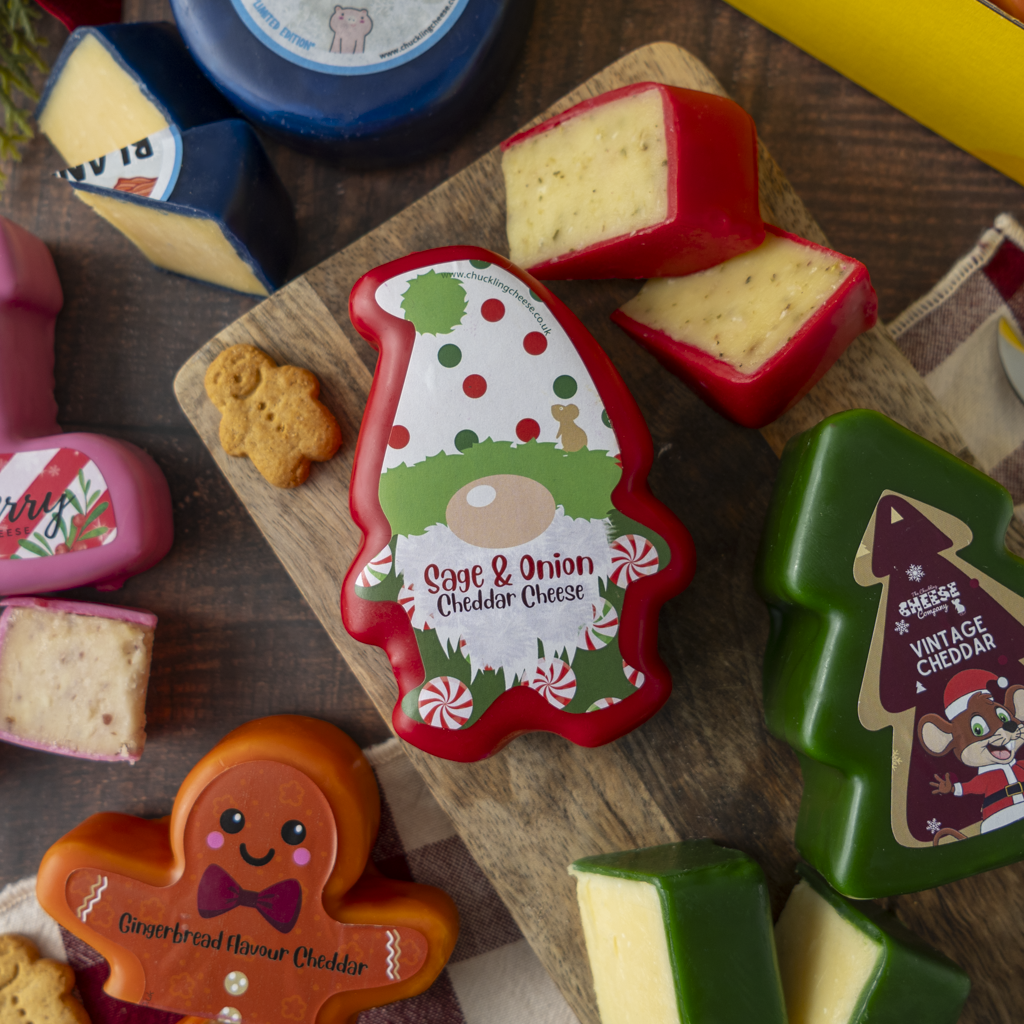 A styled cheese assortment on a wooden board, featuring a Christmas gnome-shaped cheddar cheese in the center, a gingerbread man-shaped cheddar, a green Christmas tree-shaped cheddar, and other cheese wedges and blocks around it on a dark wood surface.