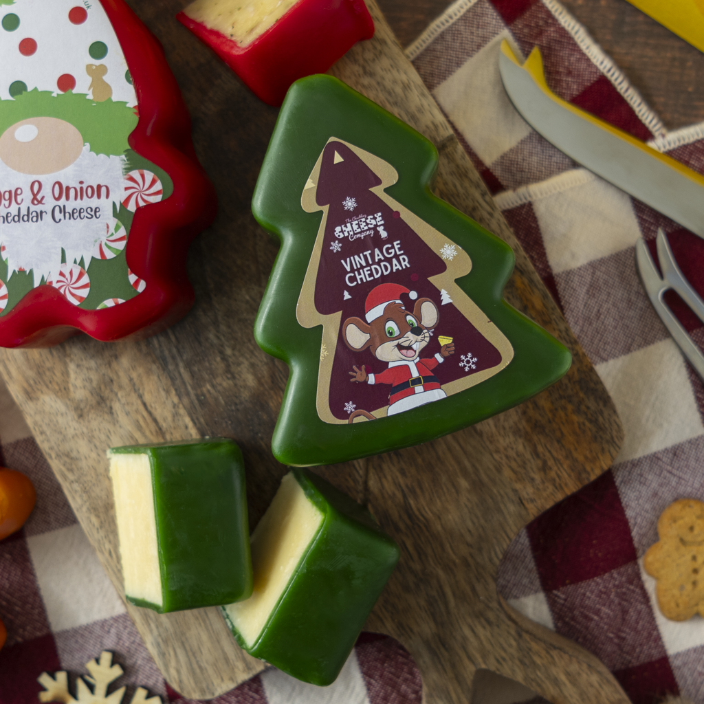 A styled product shot of Christmas-themed cheese on a wooden cutting board. The main product is a green cheese slice shaped like a Christmas tree with a label reading 'Vintage Cheddar' and a cartoon mouse in a Santa hat.