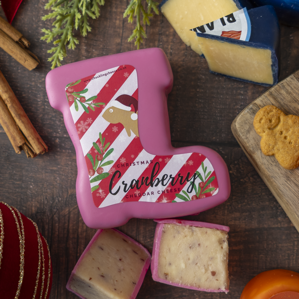 Pink Christmas boot-shaped cheddar cheese with a holiday label, surrounded by cheese wedges, cinnamon sticks, cookies, and evergreen branches on a dark wooden table.