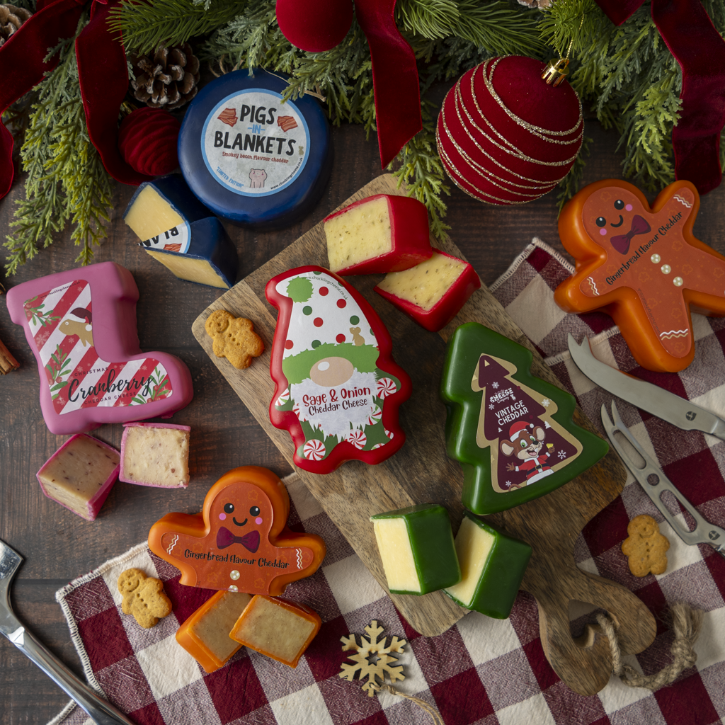 A styled holiday cheese board scene with multiple shaped cheeses and a round blue cheese labeled 'PIGS IN BLANKETS' surrounded by Christmas decorations, ribbons, ornaments, pine branches, knives, and a red-and-white check cloth.