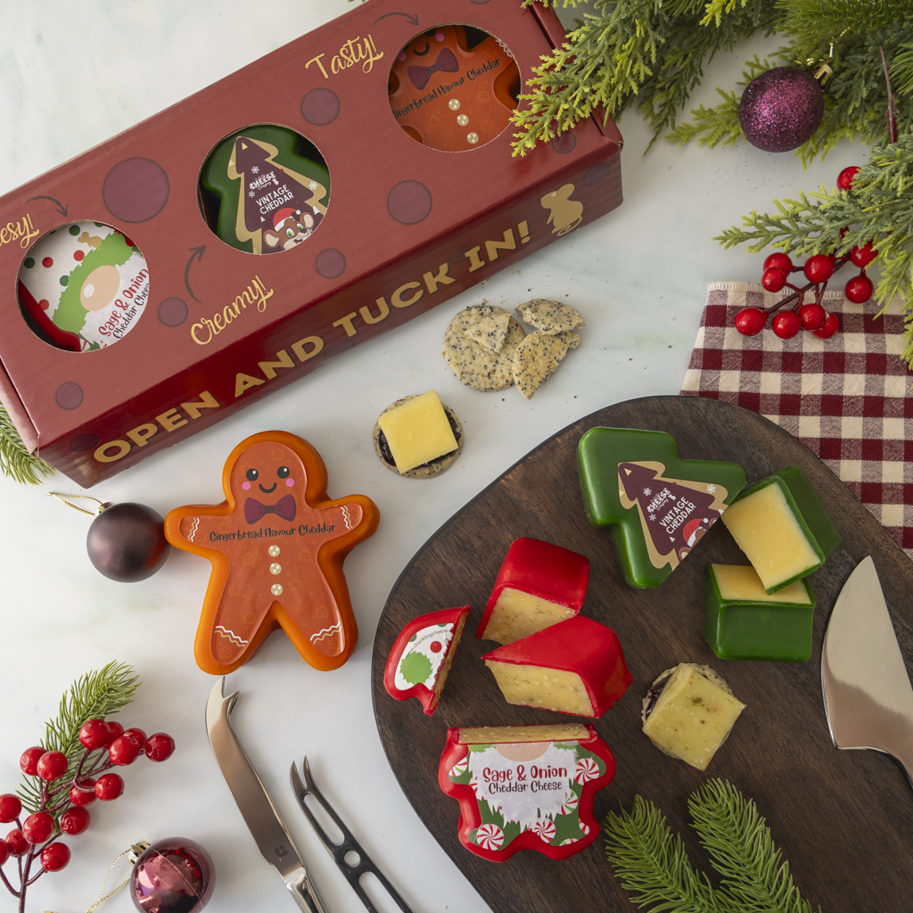 A Christmas-themed cheese and cookie charcuterie board styled on a white surface with holiday greenery, red berries, and a red gift box. The board holds several cheeses in red, green, and yellow blocks, gingerbread-shaped cookies, and crackers.