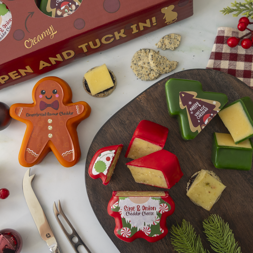 A styled holiday cheese and cookie assortment arranged on a dark wooden board with a red gift box, crackers, cheese wedges in red and green, and gingerbread-shaped cheese truckle on a white surface.
