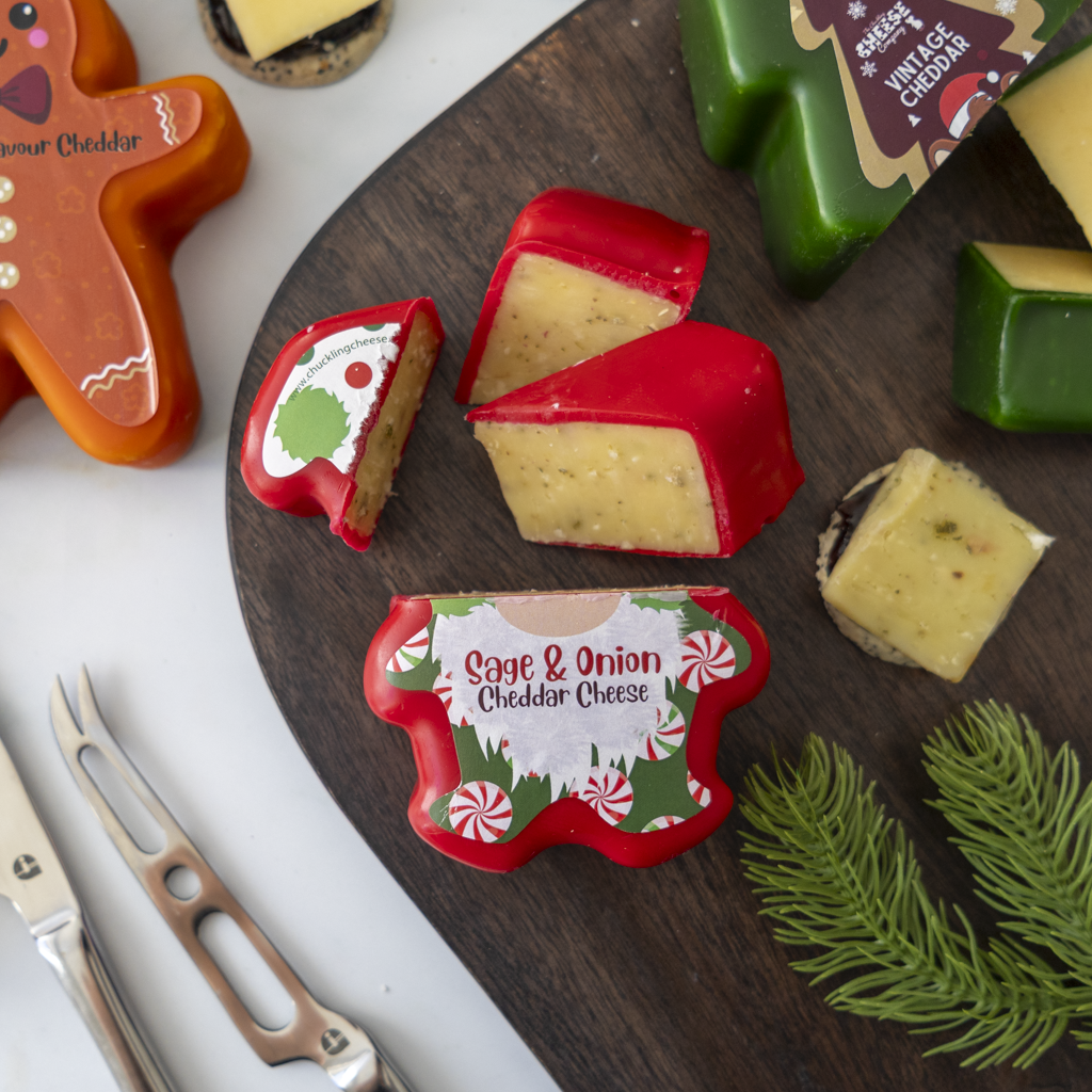 Holiday-themed cheese slices and a small cheese container with Christmas illustrations, arranged on a dark wooden board with cheese knives and other cheeses around them.