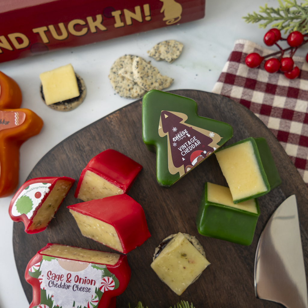 A styled holiday cheese board with several cheese wedges and slices in red, green, orange, and white holiday-themed packaging, arranged on a dark round wooden board with crackers and a knife nearby.