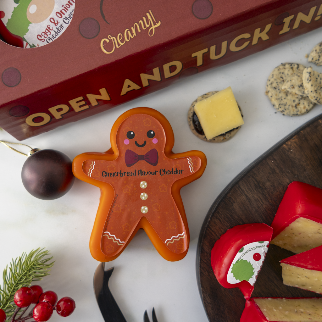 A festive Christmas-themed cheese board gift set from Sage & Onion Cheddar cheese, featuring a gingerbread man cheddar cheese truckle, a red box labelled 'Open and Tuck In!', crackers, red and yellow cheeses on a wooden board, and holiday decorations on a white surface.