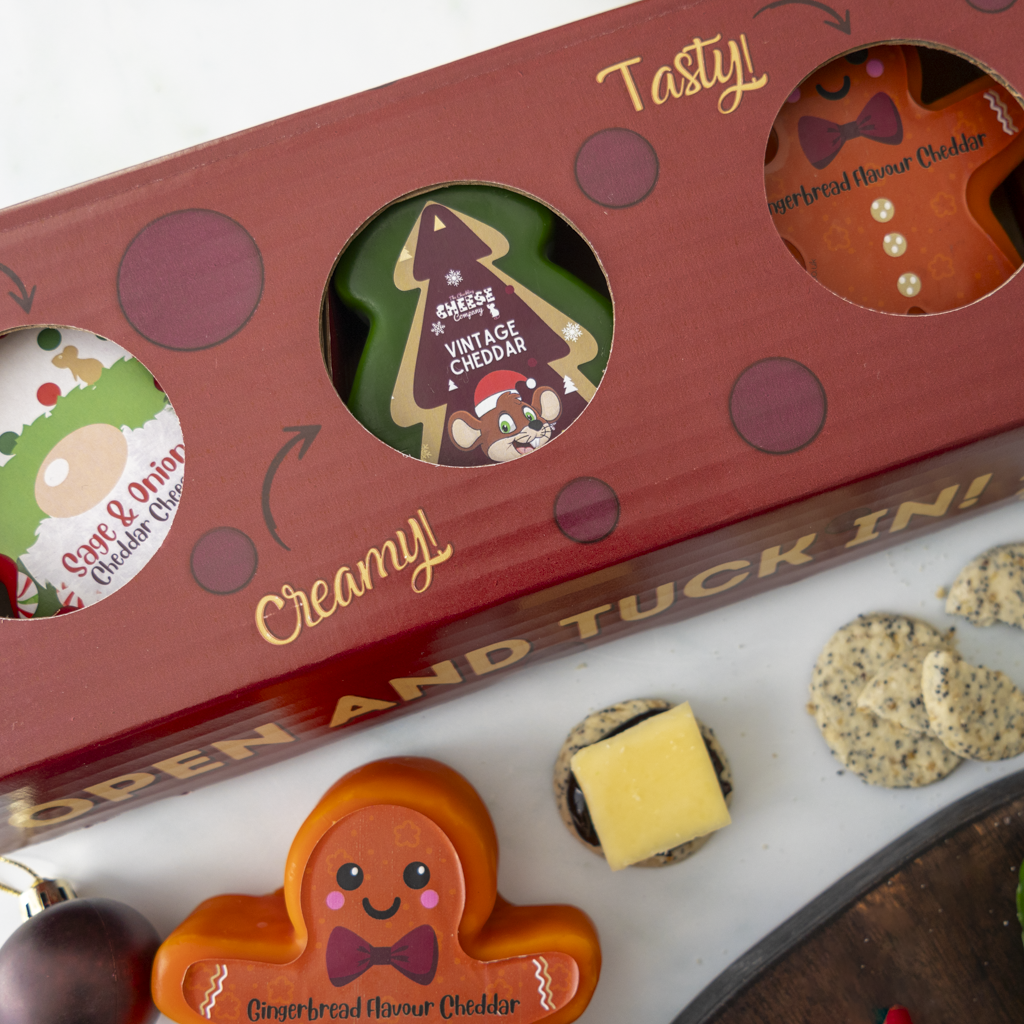 A red gift box with circular cutouts displaying three cheddar cheese products shaped like Christmas tree, gingerbread man, and a Gonk, styled on a white surface with crackers and a cheese slice nearby.