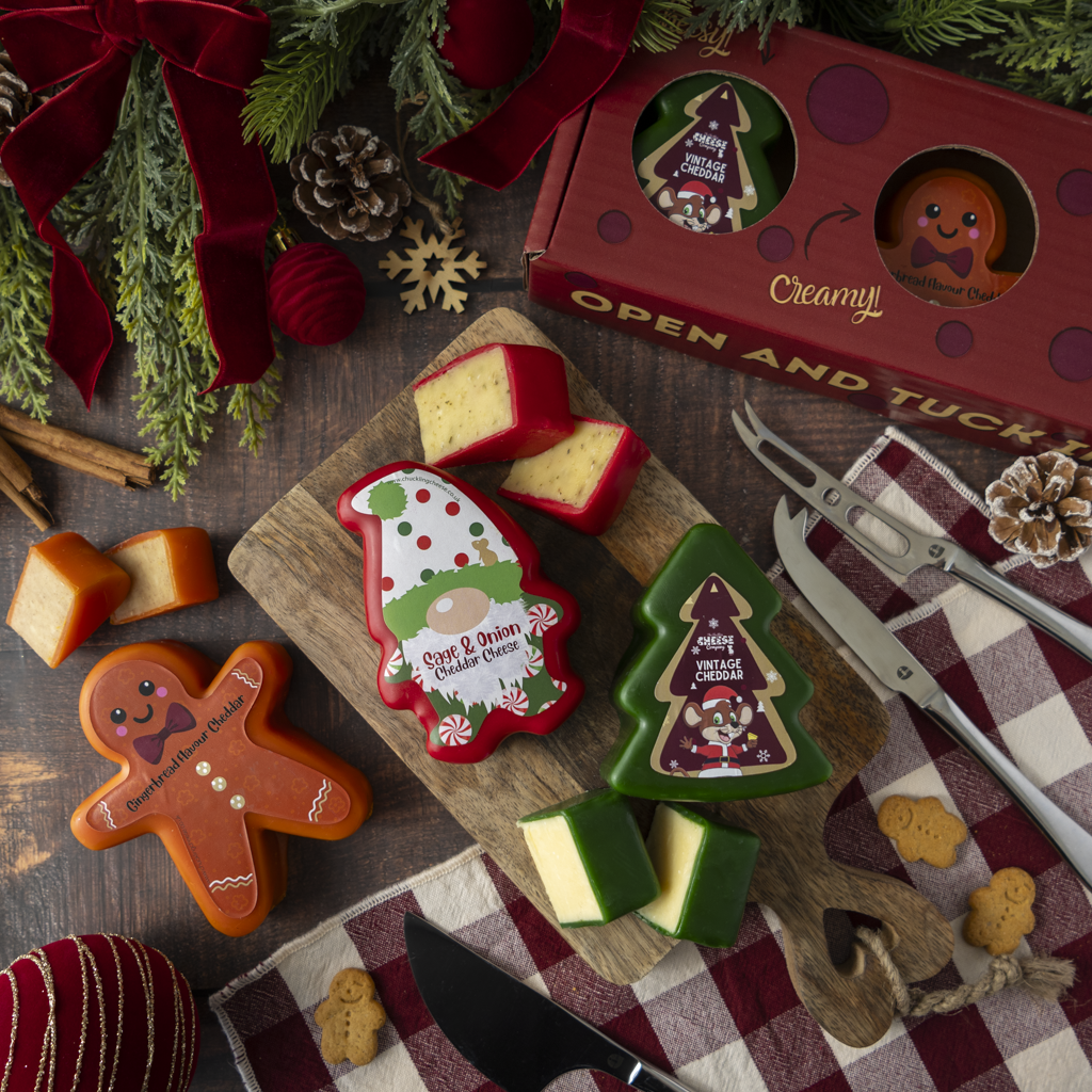 Christmas cheese truckles shaped like a gnome, a tree, and a gingerbread man, displayed on a wooden cutting board with a red gift box, cheese knives, and holiday decorations.