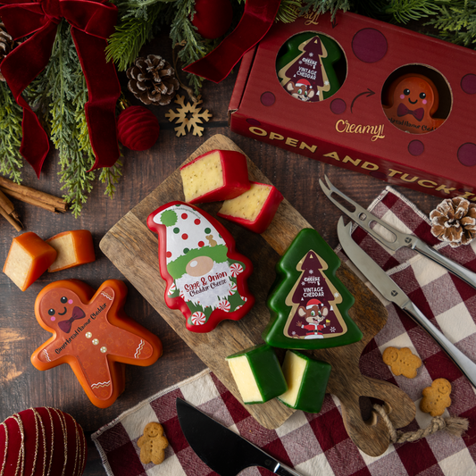 Christmas cheese truckles shaped like a gnome, a tree, and a gingerbread man, displayed on a wooden cutting board with a red gift box, cheese knives, and holiday decorations.